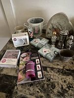 Photo of various kitchen utensils, decorative mugs, metal bucket, salt and pepper shakers, cupcake flingers, and decorative trays arranged on marble-patterned surface.