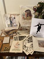 Collection of vintage advertisements and printed ephemera including color illustrated ads, black and white photos, and multiple sheets of paper with detailed drawings and prints. Displayed spread out on a surface with a faux plant in the background.