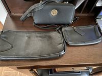 Three black purses on a wooden surface, one with a circular gold emblem, one larger plain black leather purse, and one Coach wristlet with tag and chain.