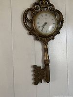 Ornate brass wall hanging clock in the shape of a key with Roman numerals on the dial, photographed against a light wall.