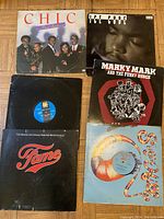 Photo of six vinyl records with covers visible on wooden floor, showing artists Chic, Ice Cube, Marky Mark and the Funky Bunch, Fame soundtrack and Sylvers.