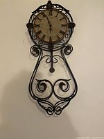 Front view of the decorative wall clock showing ornate metal frame, Roman numeral clock face, and pendulum