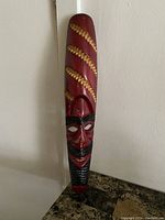 Front full view of tall carved wooden African face mask painted red with gold spiral pattern and black facial hair detailing against white corner wall.