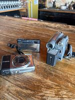 Three digital imaging devices on wooden surface - two digital cameras and one camcorder, showing their front and sides.
