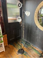 Full view of the black metal 3 light floor lamp with white lampshades, standing near a door on a wooden floor.