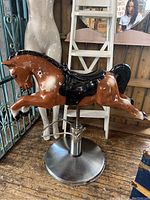 Front-side view of the horse hairdressing chair showing the brown horse figure with a black saddle and glossy finish. Mounted on a round metal base.