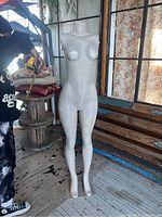 Full view of white female torso mannequin standing upright next to windowed wall and some furniture. Mannequin has no head or arms, paint is chipped all over, standing on its own but wobbly.
