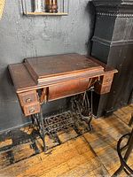 Full view of vintage New Williams sewing machine treadle table against black wall, showing feet and wooden drawers.