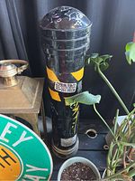 Black cylindrical parking bollard with yellow reflective tape standing near plants and a brass object. Shows wear and paint scratches.