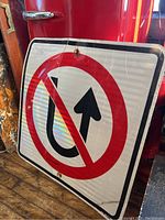 Photo of dented No U-Turn traffic sign displaying red circle and slash over a black U-turn arrow on white reflective background, leaning against surfaces.