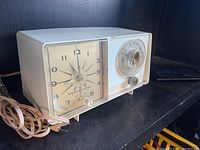 White vintage General Electric electric clock radio with analog clock and tuning dial on black shelf.
