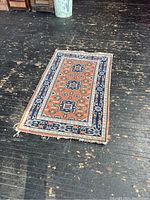 View of one vintage handwoven rug with orange field and navy blue geometric medallions, laid flat on dark wood floor. Tassels on one short edge are frayed and damaged.