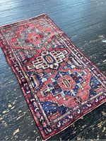 Full view of a vintage oriental rug with geometric and floral patterns in reds and blues on a dark wooden floor.