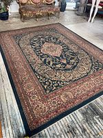 Full view of vintage rug laid flat on a wooden floor showing entire design: large central medallion, floral border, navy blue background with red and cream accents.