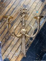 Full view of the brass wall candelabra showing two trumpet-shaped candle holders and floral ribbon details on wooden floor.