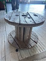 Front side view of the wooden spool table on a wooden floor, showing aged wood and caster wheels.