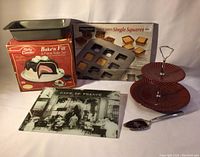 Full view of all lot items showing Betty Crocker Bake'n Fill set box, Chicago Metallic pan box, metal loaf pan, two tier red dessert dish on metal stand, metal serving spoon, and glass trivet with Cafe de France photo