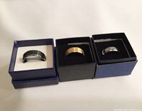 Three tungsten carbide rings in individual boxes on white background. Left ring is black, center ring is gold-tone, right ring is silver-tone.