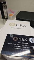 Certified moissanite gemstone displayed on a GRA Moissanite Report and warranty card showing branding and authentication.