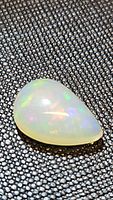 Close-up photo of a pear-shaped cabochon Ethiopian opal gemstone with visible multicolored flashes on a textured dark grey background.