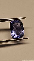 Cushion cut vibrant blue to purple natural fluorite gemstone held by tweezers against beige background, showing facets and clarity.