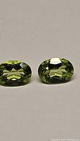 Close-up photo showing two natural apple green peridot gemstones with oval cuts placed side by side on a neutral background.