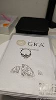 Moissanite ring placed on a GRA moissanite report booklet, showing the centered gemstone and part of the certification details.