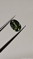 Oval cut natural green sapphire being held with tweezers showing color and clarity.