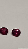 Two oval cut vivid blood red rubies shown side by side on a neutral background with visible facets and deep red color.