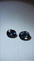 Pair of oval cut teal blue sapphires displayed on a flat surface with direct lighting to show color and clarity.