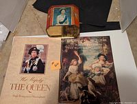 Photo showing two books about Her Majesty The Queen and The Queen's Pictures along with a vintage biscuit tin featuring the Queen's photo.