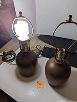 Two teak wood table lamps placed on white surface, one fitted with lit bulb, showing wood grain and brass socket detail.