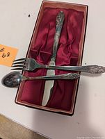 Three-piece silver plated cutlery set (fork, knife, spoon) displayed inside original red satin-lined box.