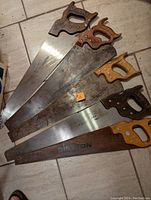 Five hand saws laid out on a tile floor showing variety in handle shapes and blade conditions.