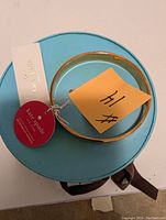 Photo of the gold-tone Kate Spade bangle bracelet, showing the front and top view with original tag attached, placed on blue circular box with brown ribbon.