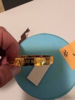 Photo of gold-tone Kate Spade bangle bracelet showing the phrase 'Play Nice' engraved on it held over a blue round box.