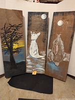 Three painted barn boards displayed vertically side by side: tree at sunset, howling wolf, and bunny looking up at moon.