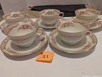 Five Noritake cups paired with matching saucers arranged in two rows on a table, white base with floral and gold accents.