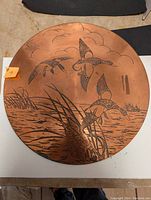 Front of the round copper plaque showing etched scene of three flying ducks over marshland reeds and water. Copper surface is bright with a warm tone and some patina.