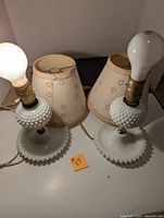 Pair of white milk glass lamps with hobnail pattern and beige floral fabric shades, bulbs installed, placed on a white surface.