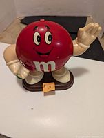 Front view of the red M&M character dispenser showing the face, white gloves, and base