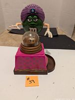 Front view of the green M&M fortune teller candy dispenser showing the character and the pink decorated base with clear dome.
