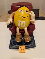 Front view of Yellow M&M character seated in red recliner holding remote on blue rug, showing details and facial expression.