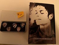 Photo showing Michael Jackson Unauthorized book with a serene close-up portrait on the cover alongside the metal tin containing 5 guitar picks with various images of Michael Jackson.