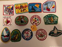Full view of 13 colorful embroidered scout badges from 1975 to 2001 with various camp and scouting themes including animal mascots and event logos.