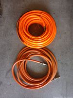 Two coiled orange machine air hoses, one looking newer and cleaner, the other with some marks and oily residue, set on a concrete floor.