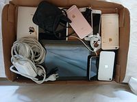Box containing several Apple iPhones in various colors and models, black iPad, and tangled white charging cables and chargers.