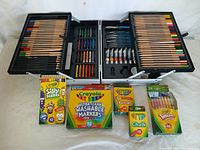 Black case open showing colored pencils, oil pastels, graphite pencils, and paint tubes. Boxes of Crayola Silly Scents crayons, washable markers, 24-count crayons, chalk, and Twistables colored pencils arranged in front of case.