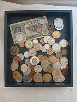 Top-down view of assorted foreign coins and one banknote in a black tray, showing coins of different sizes and materials, including some with holes and bi-metallic designs, alongside a Netherlands 2½ Gulden banknote.
