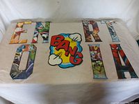 Set of eight metal superhero letters arranged on a flat surface with colorful comic 'BANG' graphic piece centered among them.